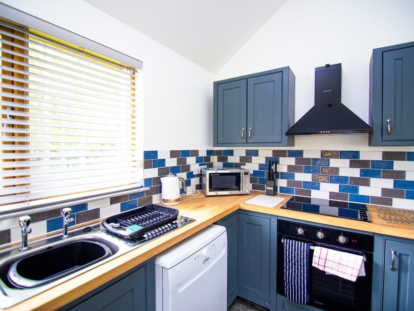 A compact kitchen with a dishwasher, microwave, and oven, featuring blue cabinets and a tiled backsplash.