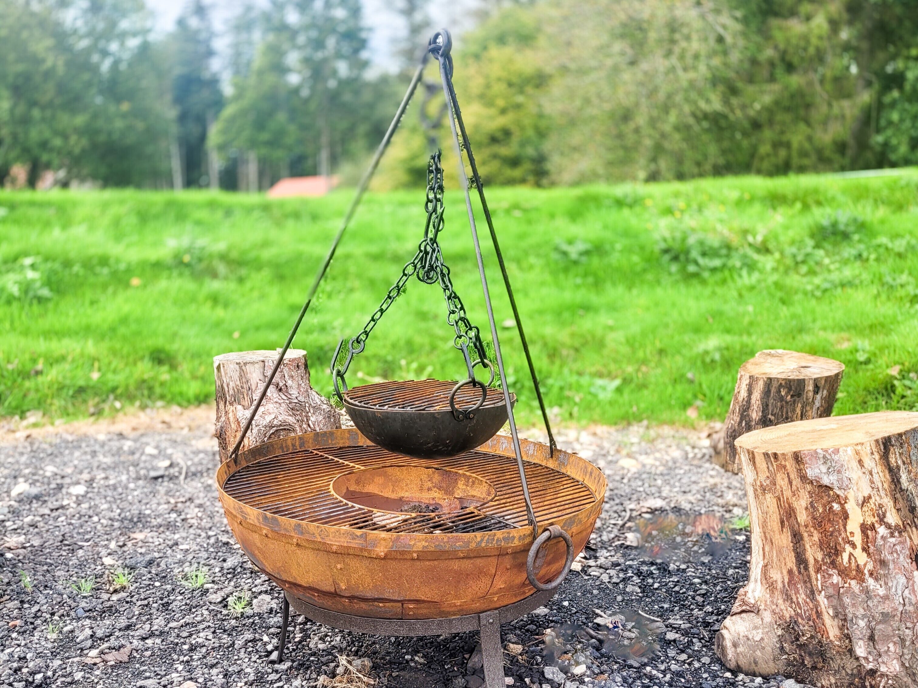 Una parrilla rústica al aire libre con un recipiente metálico suspendido sobre ella, sostenido por cadenas y un soporte metálico, ubicada sobre un terreno de grava con dos troncos de madera cerca.