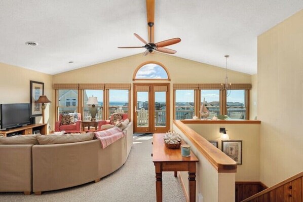 A spacious living room with a large window, a ceiling fan, and a television. - OceanSound - 500 Feet to the Ocean in Duck, NC! (Kitty Hawk)