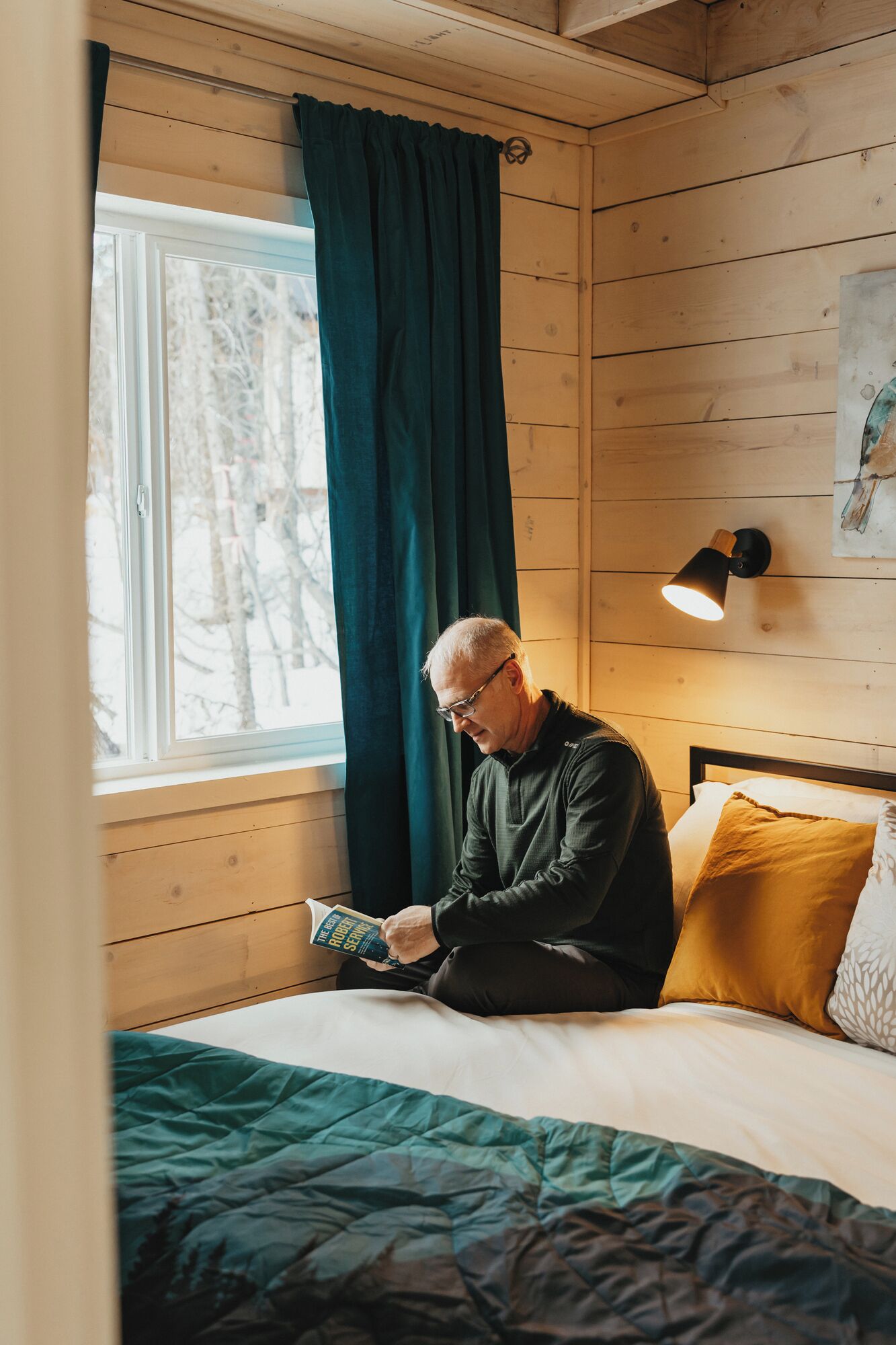 A person sitting on a bed in a room with wooden walls, reading a book.