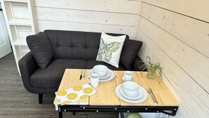 A dining area with a wooden table set for two, a gray sofa, and a green armchair. A framed painting of a mountain landscape hangs on the wall.