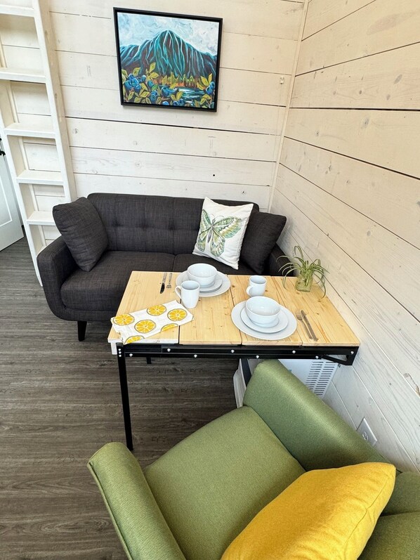 Un comedor con una mesa de madera dispuesta para dos personas, un sofá gris y un sillón verde. Un cuadro enmarco de un paisaje montañoso cuelga en la pared.