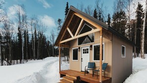 Een houten hut met een veranda en twee stoelen, omgeven door sneeuw en bomen.