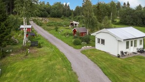 Een landelijk gelegen bezit met een grindpad dat leidt naar een wit huis met een groot raam en een veranda. Er zijn meerdere rode gebouwen met uiteenlopende daken, omringd door weelderig groen en bomen.