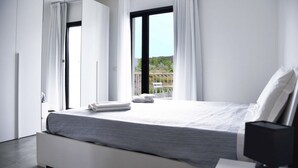 A modern bedroom with a large bed, white curtains, and a view of a balcony with outdoor furniture.