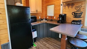 A compact kitchen with wooden cabinets, a black refrigerator, a coffee station, and a small bar area.