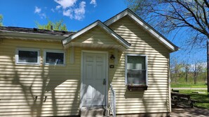 A small, single-story house with a white door, two windows, and a small porch area.