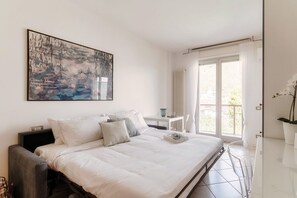 A bedroom with a bed, a nightstand, a table, a chair, a window with curtains, and a painting on the wall.