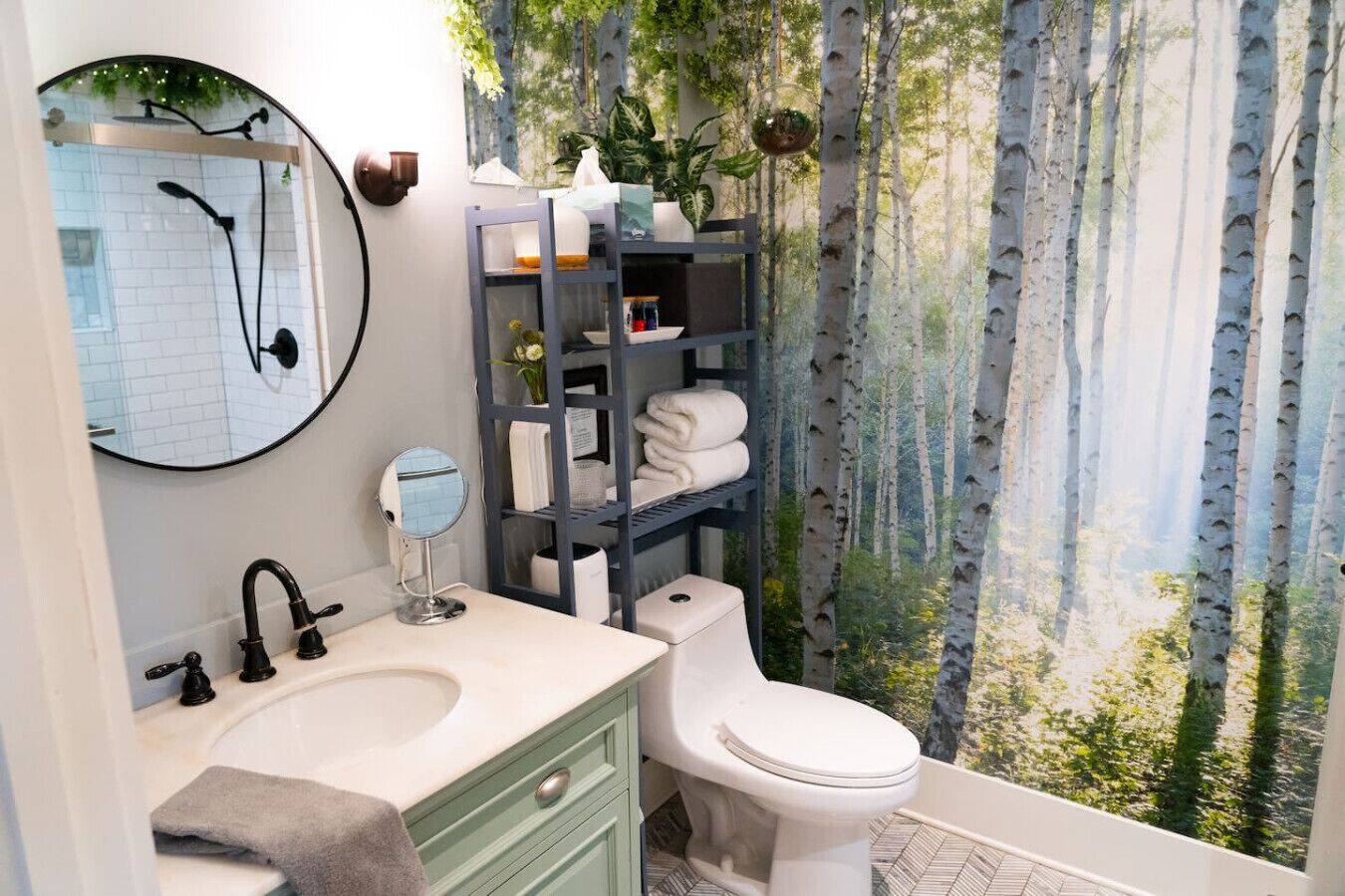 Ein Badezimmer mit Waschbecken, schwarzen Wasserhähnen, einem runden Spiegel, einer Toilette und einem Regal mit Handtüchern.