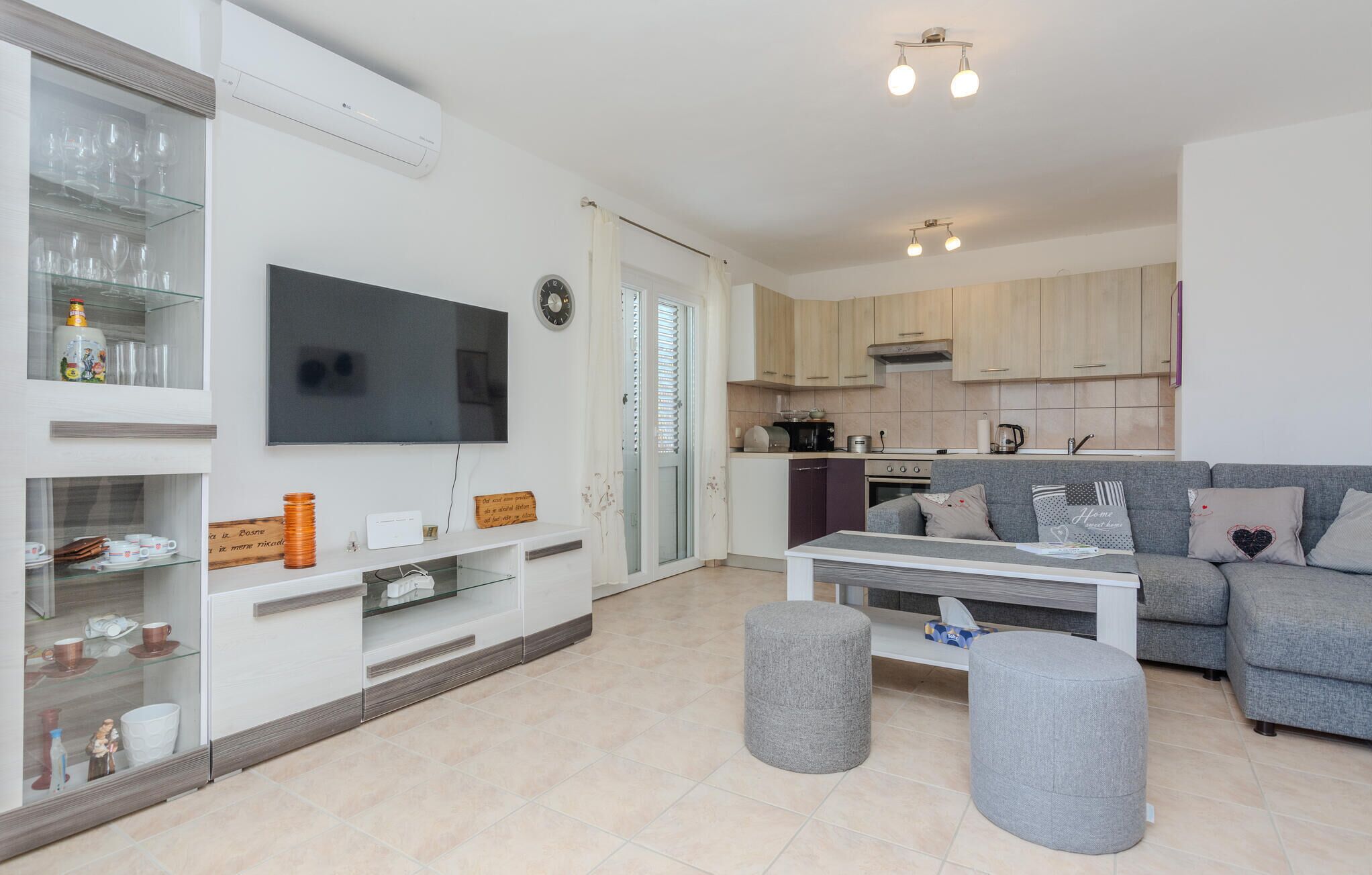 A modern living room with a flat-screen TV, a grey sofa, ottomans, and a kitchen area with wooden cabinets and a microwave.