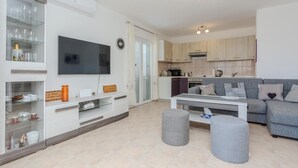 A modern living room with a flat-screen TV, a grey sofa, ottomans, and a kitchen area with wooden cabinets and a microwave. - Nice home in Privlaka (Privlaka)