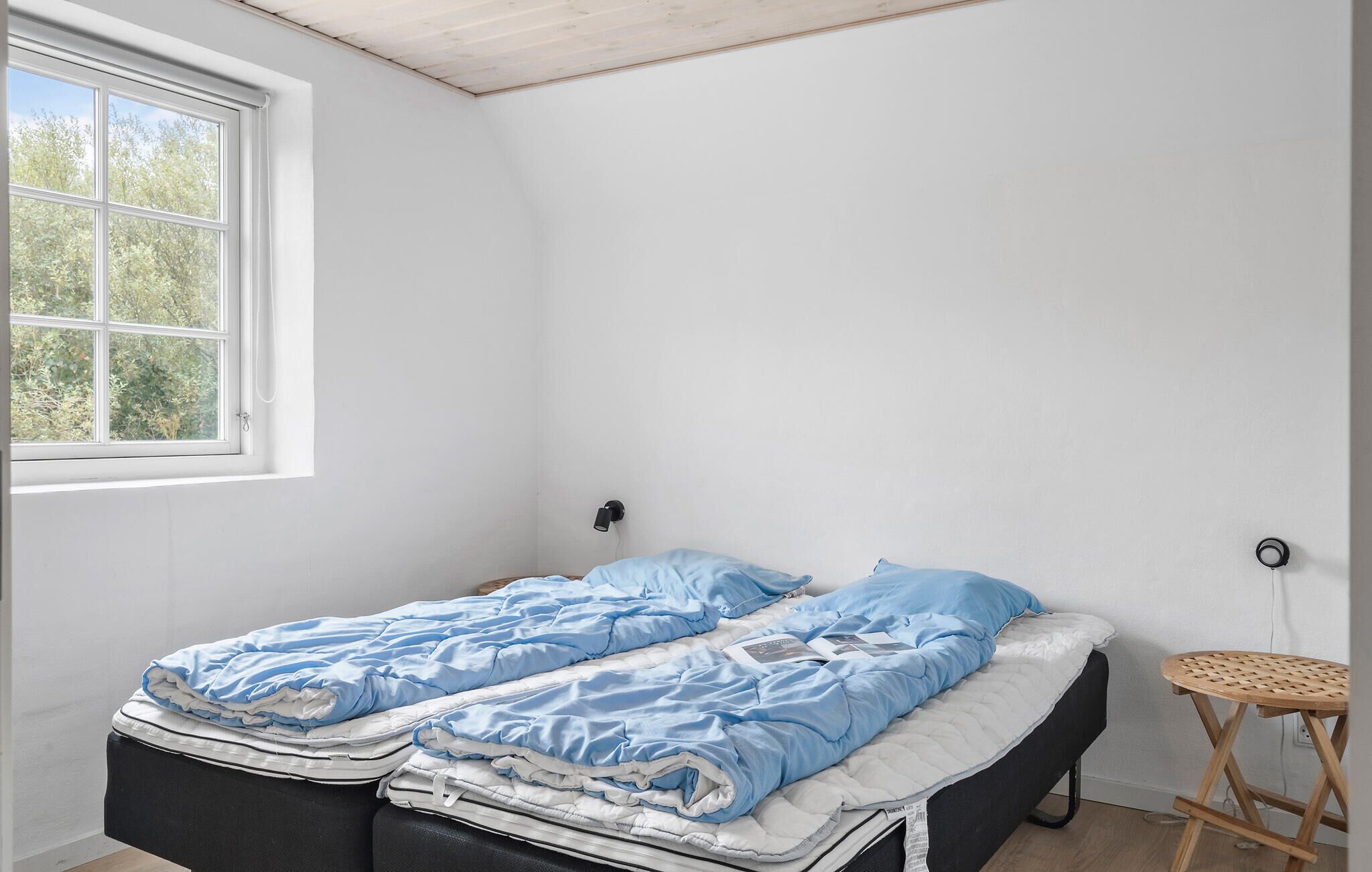 Une petite chambre avec un lit simple, un plafond en bois, une fenêtre donnant sur les arbres, un petit tabouret en bois et une lampe fixée au mur.