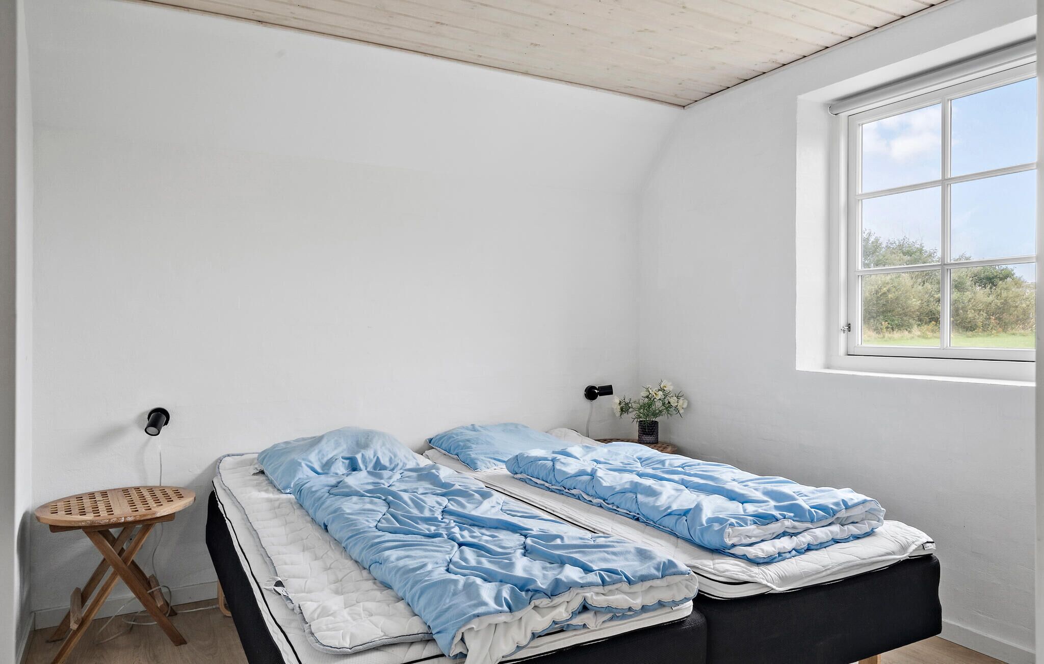 A bedroom with a bed, a small wooden stool, a potted plant, and a window with a view of greenery.