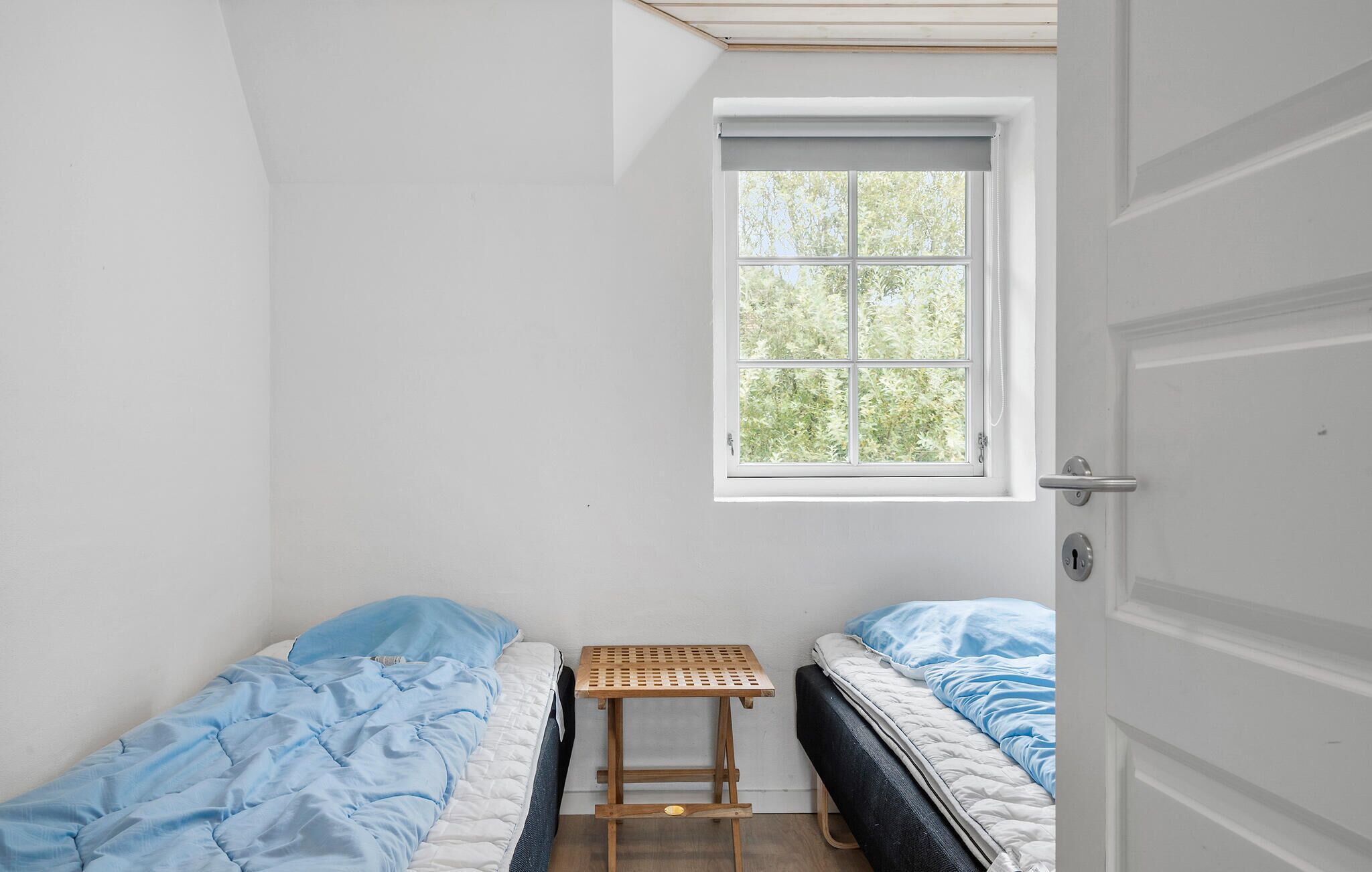 A small, single-bed room with a window, a wooden stool, and a door.