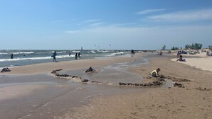 En strand där folk promenerar och leker i vattnet, ett fyrtorn i fjärran och en sandstrand med några stenar.