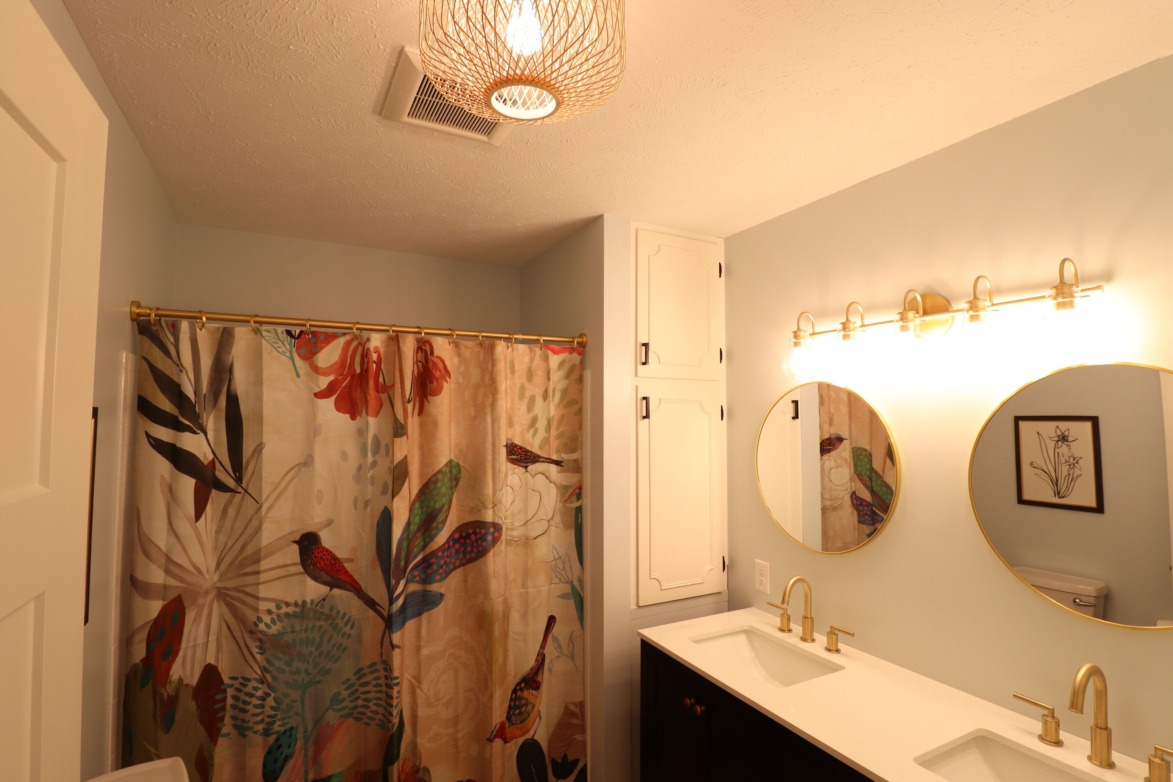 A bathroom with a shower curtain featuring a bird and leaf pattern, a white sink with gold fixtures, and two round mirrors with gold frames.