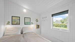 Une chambre à coucher avec un lit, deux tableaux encadrés, une petite étagère avec un vase et une fenêtre donnant sur un paysage verdoyant.