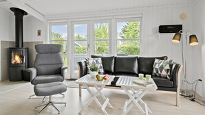 Un salon moderne avec un poêle à bois noir, un fauteuil gris, un canapé en cuir noir et une table basse blanche agrémentée de livres et de fruits.