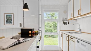 Une cuisine moderne avec des meubles blancs, une grande fenêtre et un coin repas où l’on aperçoit un livre et des pommes sur le comptoir.