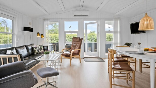 Une pièce spacieuse avec un canapé en cuir noir, un fauteuil gris, une table à manger en bois avec des chaises, et une grande fenêtre donnant sur un paysage verdoyant.