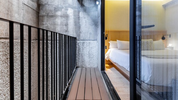 Una habitación de hotel moderna con pared de concreto, piso de madera, cama con sábanas blancas y separador de vidrio.