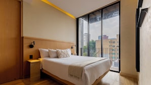 Un dormitorio moderno con una cama grande, cabecero de madera, mesita de noche y vistas a la ciudad a través de un ventanal.