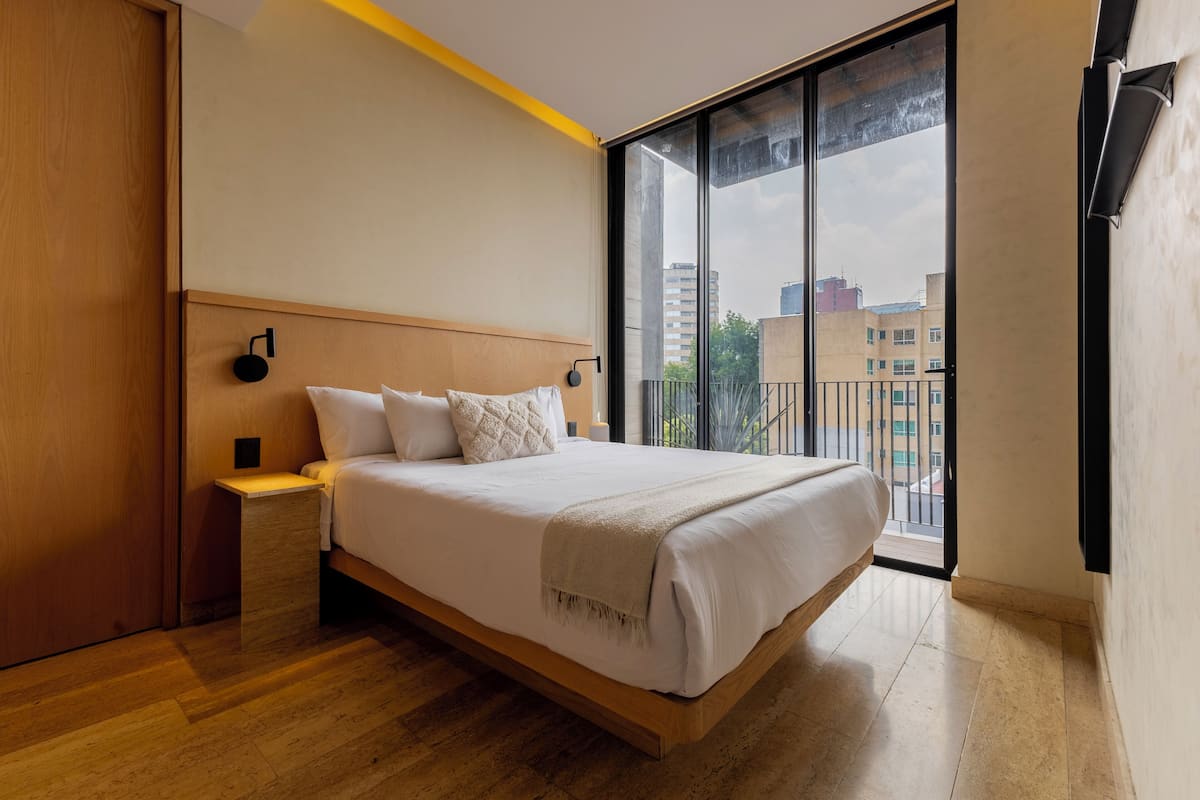 Une chambre moderne avec un grand lit, une tête de lit en bois, une table de chevet et une vue sur la ville grâce à une grande fenêtre.