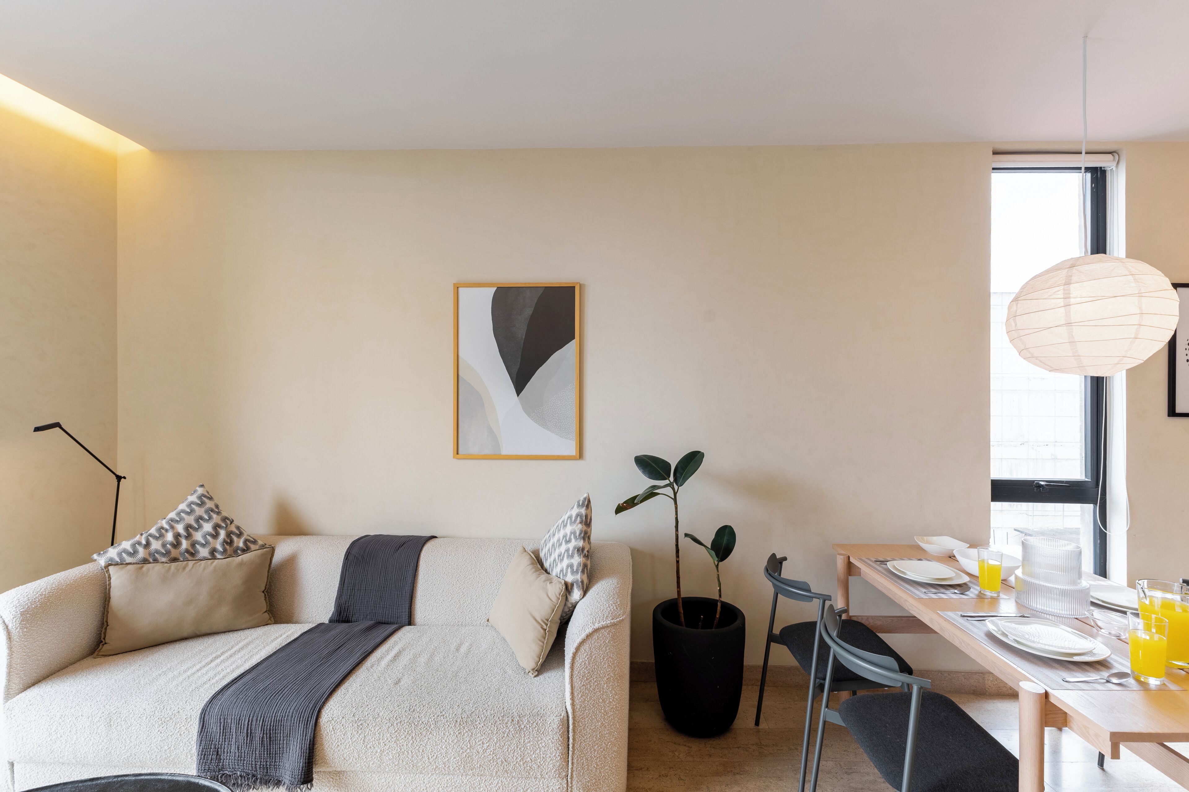 A modern living room with a sofa, a dining table set for breakfast, a potted plant, and a framed abstract artwork on the wall.