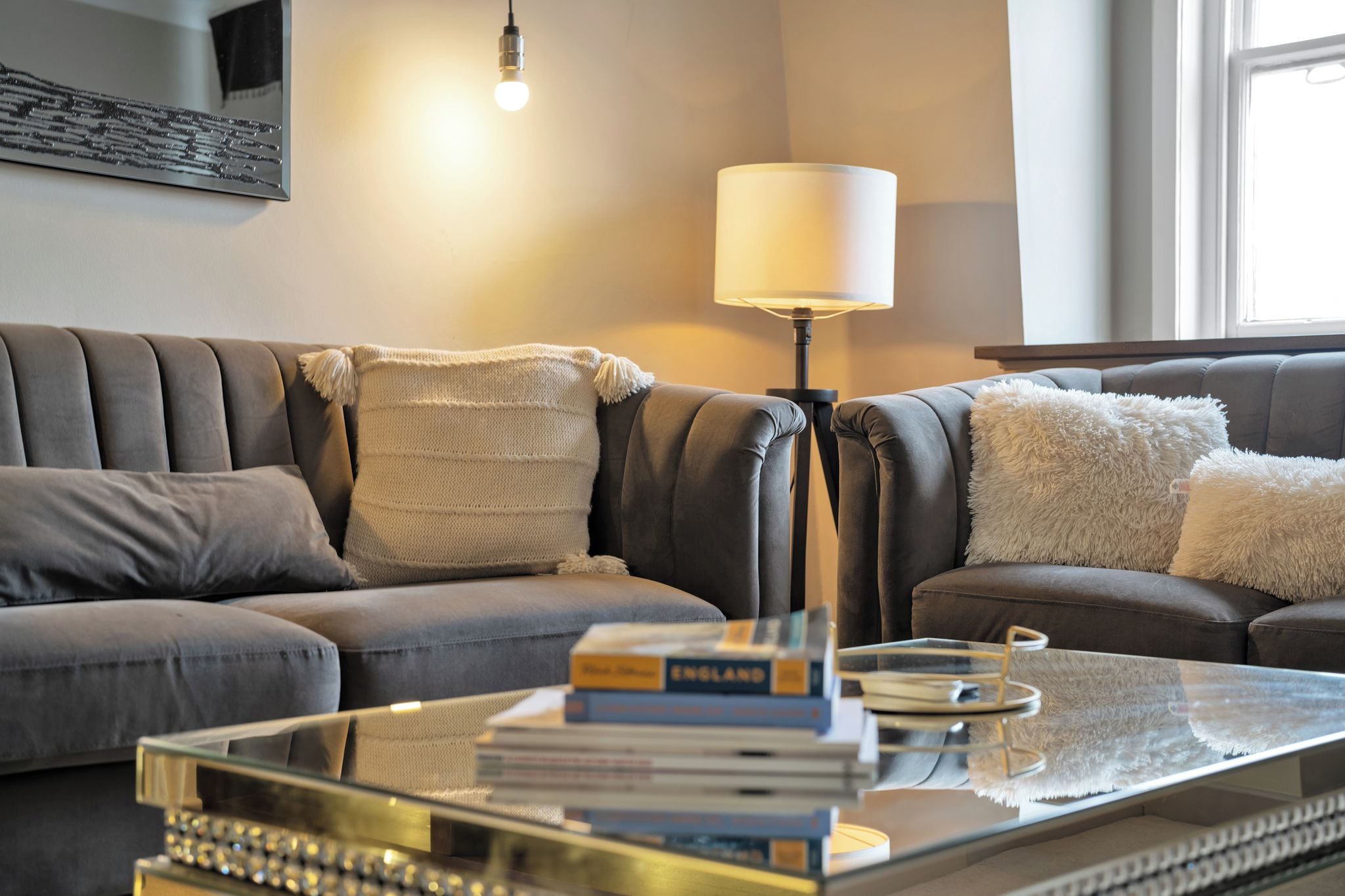 Una sala de estar moderna con un sofá gris, una mesa de centro de vidrio con libros, una lámpara de pie y un ventanal con luz natural.