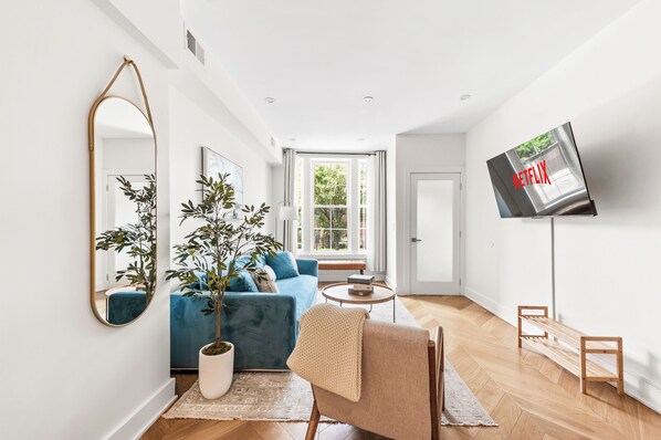 A modern living room with a blue sofa, a potted plant, a wooden bench, and a flat-screen TV mounted on the wall. - 3 LEVEL LUX Townhome! Near MetLife NYC EWR PATH, 4Bed/3Bath! (Jersey City)