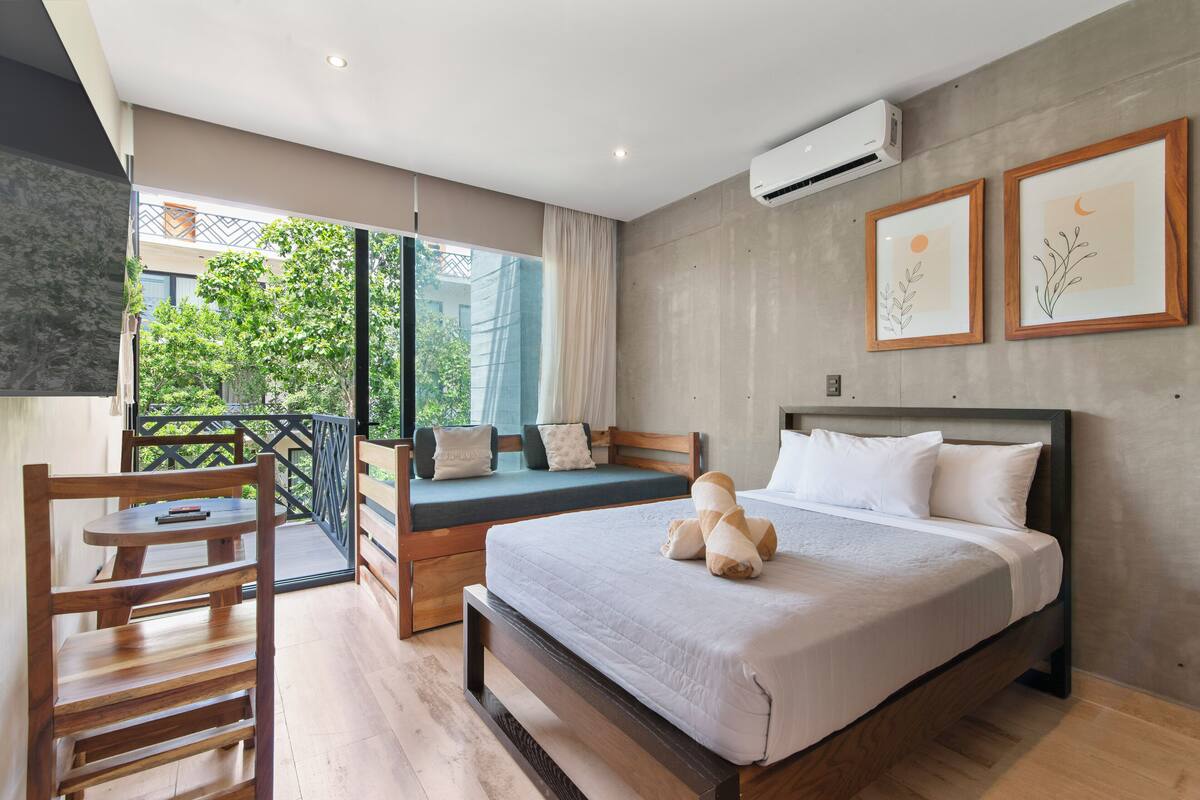 Ein modernes Schlafzimmer mit einem Bett, einem Holzsessel, einem Balkon mit Blick auf Grünflächen und zwei gerahmten Bildern an der Wand.