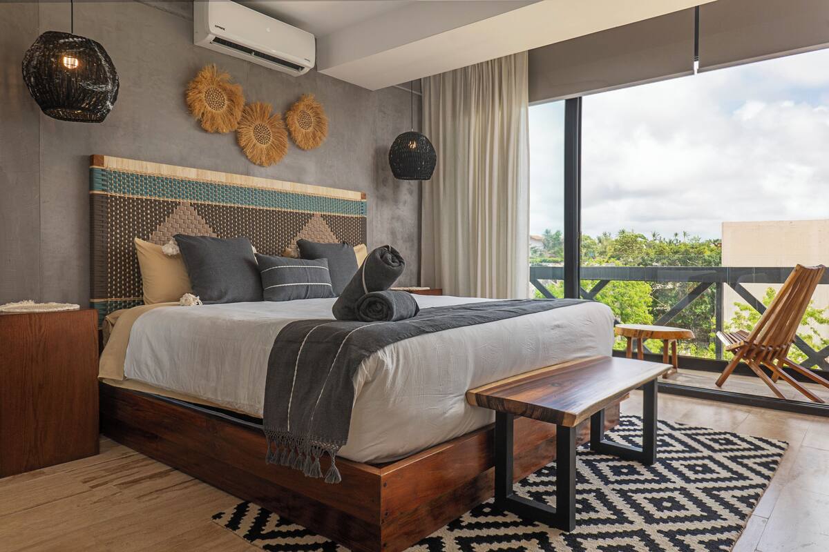 Ein modernes Schlafzimmer mit einem großen Bett, einer hölzernen Sitzbank, Blick ins Grüne und einem Balkon mit einem Stuhl.