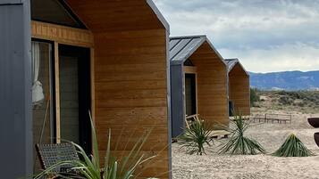 A row of modern, wooden cabins with large sliding doors, set in a desert landscape with sparse vegetation and distant mountains.