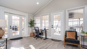A spacious room with a desk, office chair, and a comfortable chair. There are large windows with blinds, a potted plant, and a small table with a lamp. - Lakefront Getaway: Fire Pit ~ Dock ~ Games ~ Family & Pet Friendly (Waterloo)