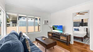A living room with a sofa, coffee table, TV, and a bed with a ceiling fan. - NEW Private Ocean Beach Home with Yard and Parking (San Diego)