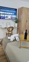 Habitación de hotel ordenada con cama, un armario de madera, una mesita con una botella y vasos, una silla, un estante con una planta y un televisor mostrando la interfaz de un servicio de streaming.