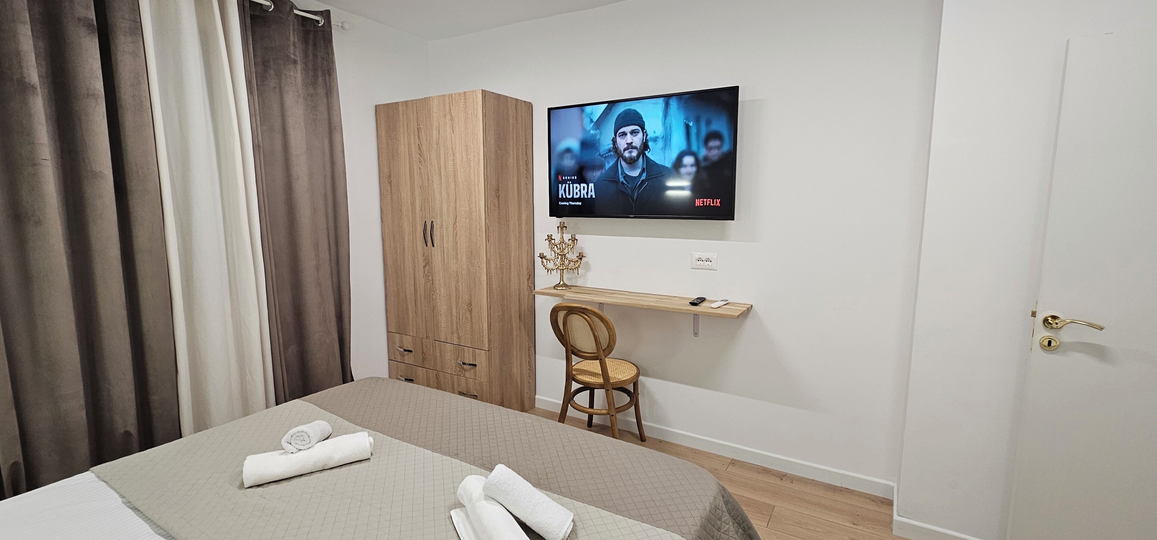 A hotel room with a bed, a TV mounted on the wall, a wooden wardrobe, a desk with a chair, and a door.