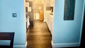 Een smalle gang die uitkomt bij een keuken met witte kastjes, een houten vloer en een plafond met inbouwverlichting.
