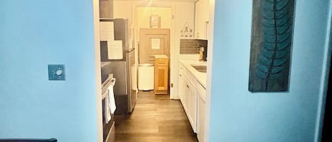 A narrow hallway leading to a kitchen with white cabinets, a wooden floor, and a ceiling with recessed lighting.