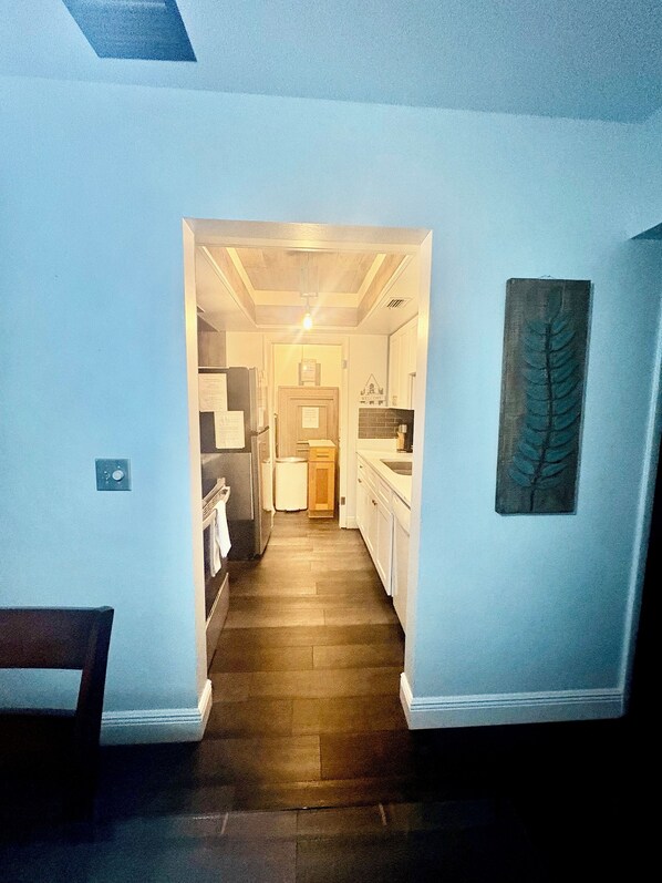 A narrow hallway leading to a kitchen with white cabinets, a wooden floor, and a ceiling with recessed lighting.