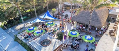 An outdoor area with a thatched-roof structure, multiple umbrellas, and people gathered around tables and chairs.