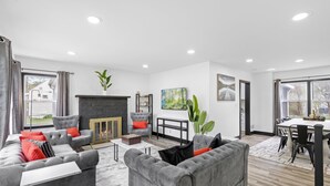 A modern living room with grey sofas, a fireplace, a dining area, and large windows. - Roomy Retreat–Family&Friends|2 King Beds|Game Room | Valpo University | Dunes (Chesterton)