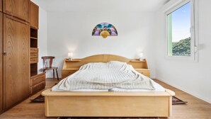 Ein Schlafzimmer mit einem hölzernen Bett, Nachttischen mit Lampen, einem Holzschrank und einem Fenster mit Blick auf Grünflächen.