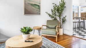Un salon avec un plancher en bois, une table basse ronde en bois sur laquelle est posée une plante, un fauteuil vert, une plante en pot et un tableau représentant une scène de plage.