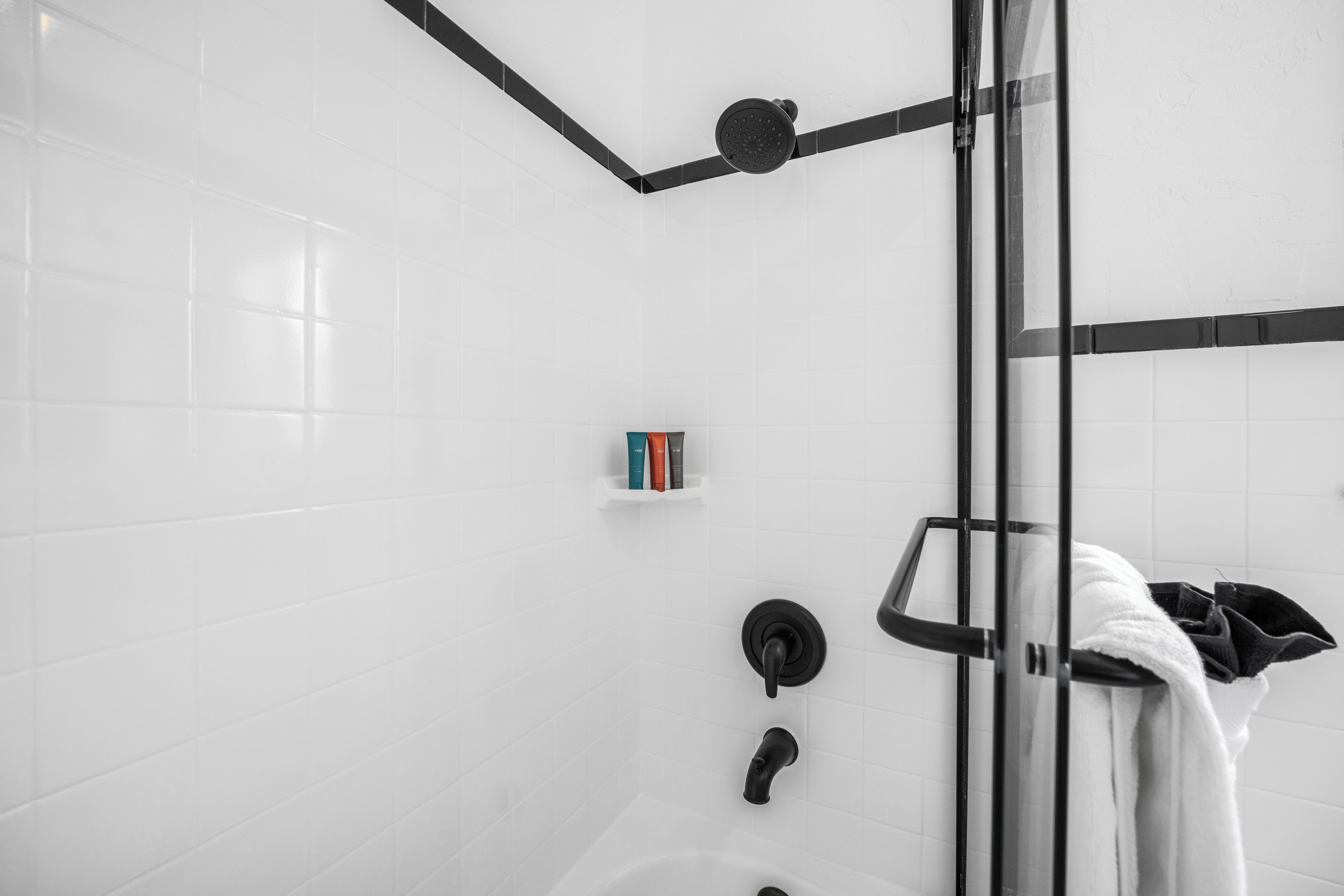 A modern bathroom with white tiles, a glass shower enclosure, a towel rack, and a small shelf with books.
