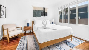 Une chambre à coucher comprenant un lit, une chaise, une table de chevet, une décoration murale en macramé et une fenêtre avec des stores.