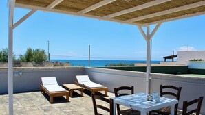 Ein überdachter Außenbereich mit Tisch und Stühlen, zwei Liegestühlen und Blick auf das Meer.