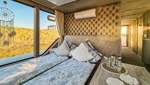 Une chambre avec deux lits, une fenêtre donnant sur un paysage désertique, un climatiseur fixé au mur et une table avec un set de table et un pot.