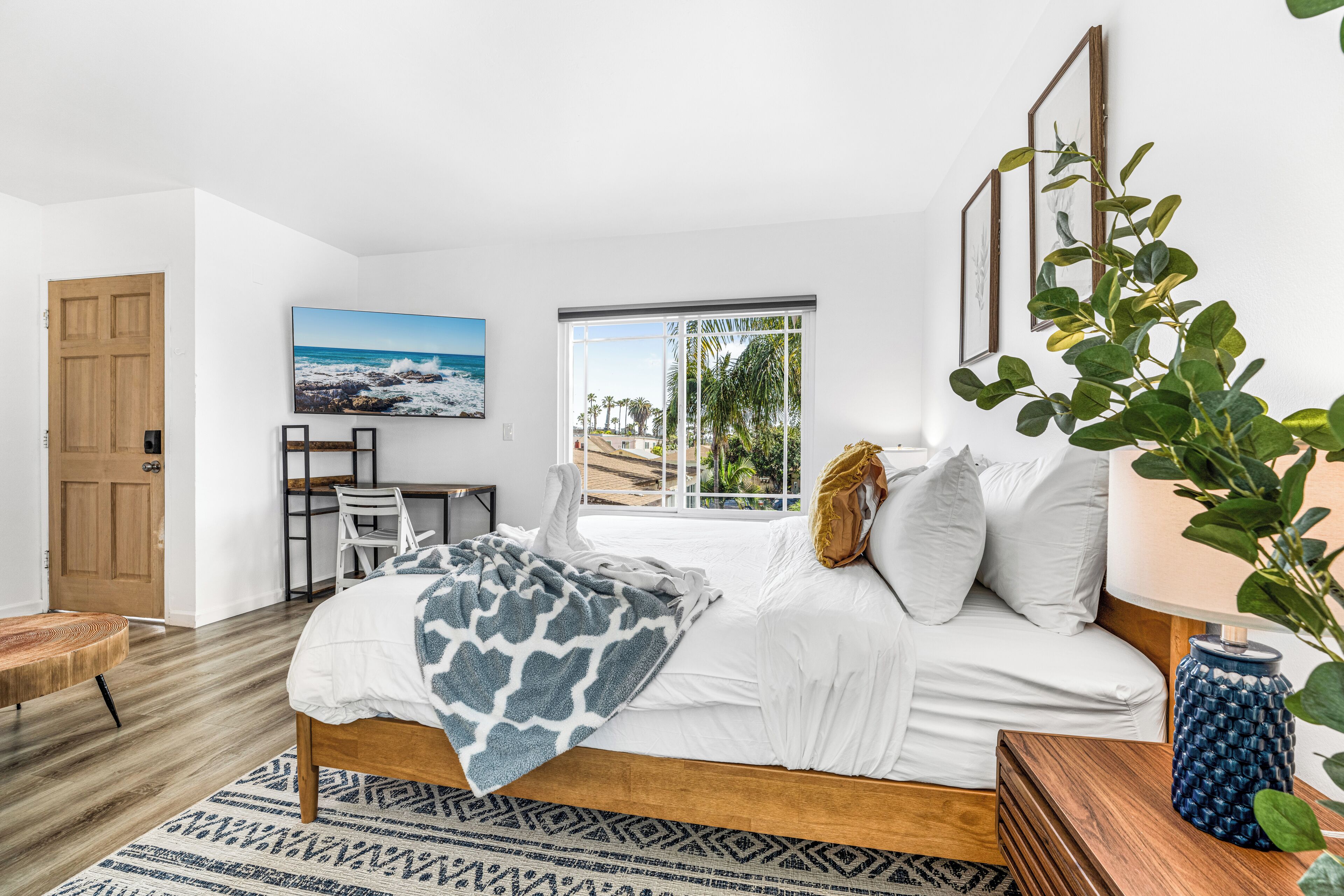 A bedroom with a bed, a desk, a chair, and a large window with a view of the beach.