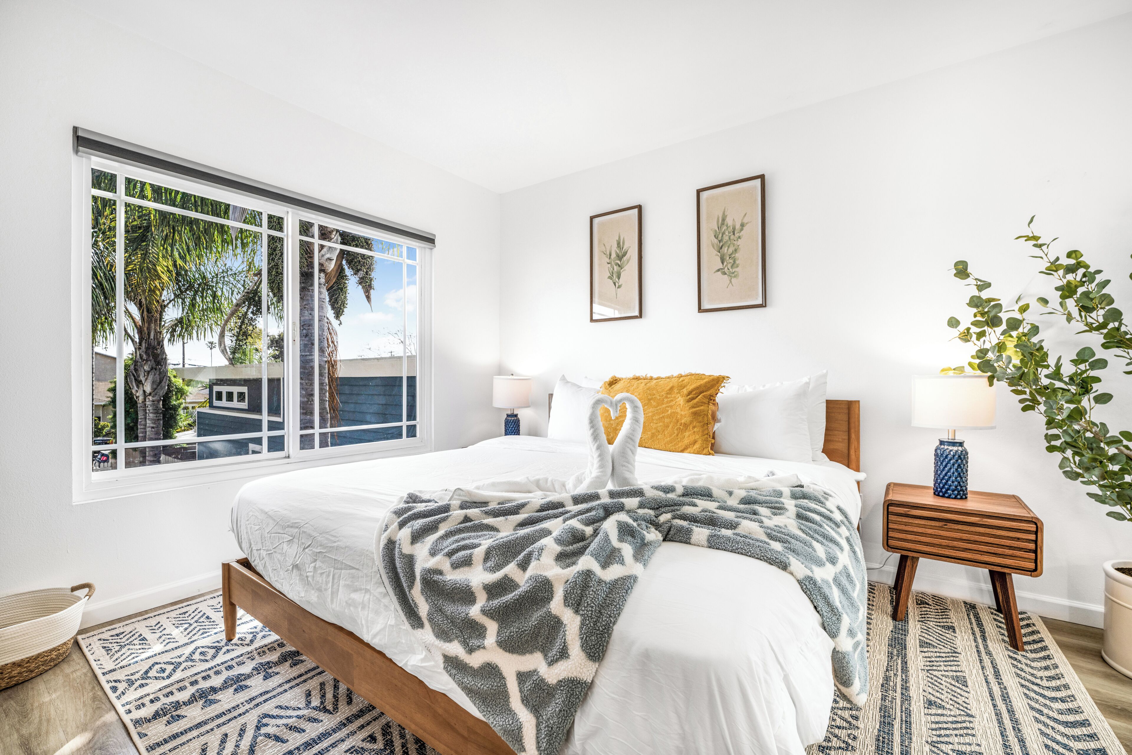Une chambre avec une grande fenêtre, un lit recouvert d’une couverture à motifs, une table de chevet avec une lampe et un tableau accroché au mur.